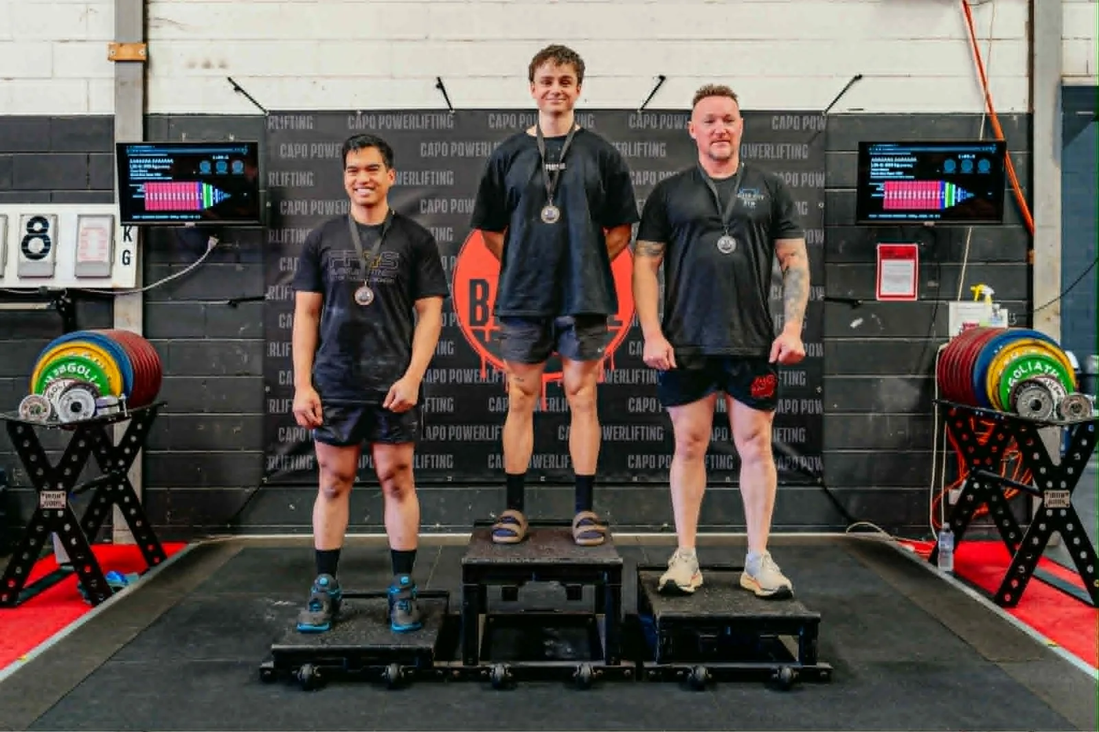 Three powerlifters on the podium at a CAPO sanctioned meet, medals around their necks, including a Massages By Sarah-supported lifter.