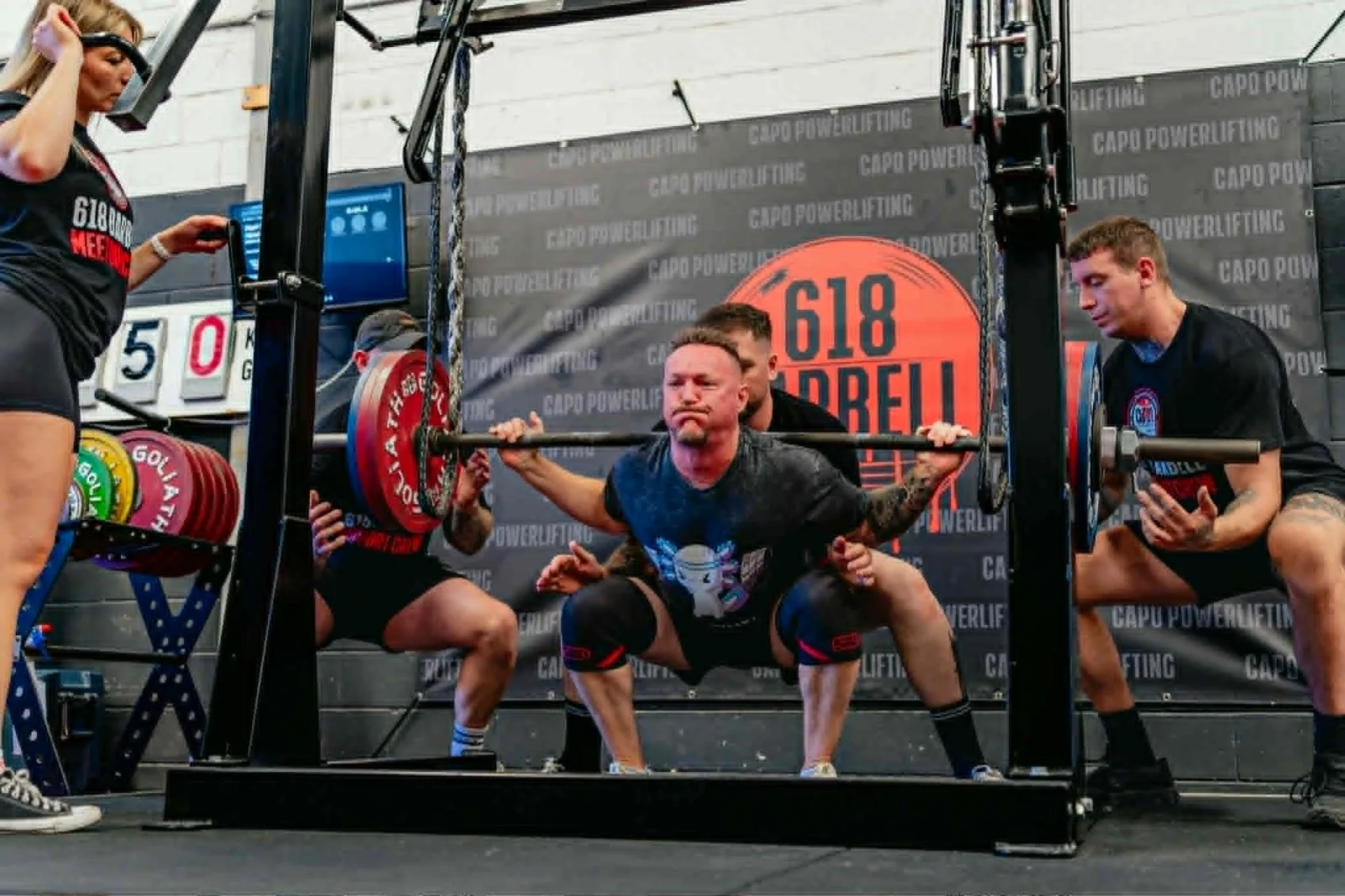 Nigel deep in a heavy back squat at a CAPO sanctioned powerlifting meet with spotters either side and a chain-loaded barbell across his back.